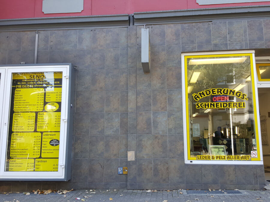 Blick auf die gelb-schwarz gehaltene Schaufenstergestaltung zweier Lokale, einer Imbissstube und einer Änderungsschneiderei mit freier Hauswand dazwischen.